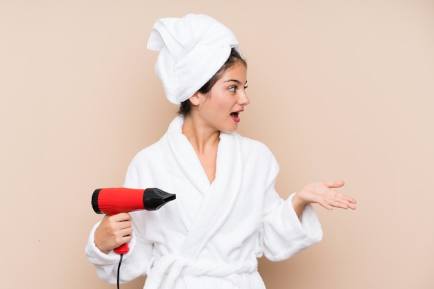 Jovem mulher em um roupão de banho com secador de cabelo mais isolado com expressão facial de surpresa