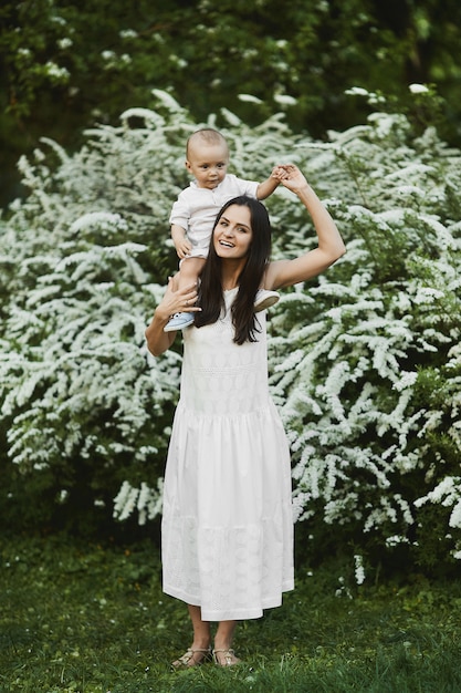 Jovem mulher em um vestido longo branco com um bebezinho bonitinho na camisa e shorts andando no jardim verdejante em dia de verão