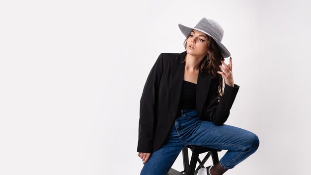 Jovem mulher encantadora com casaco preto e chapéu cinza sentada em uma pequena escada de madeira e olhando para a câmera isolada sobre um fundo cinza do estúdio