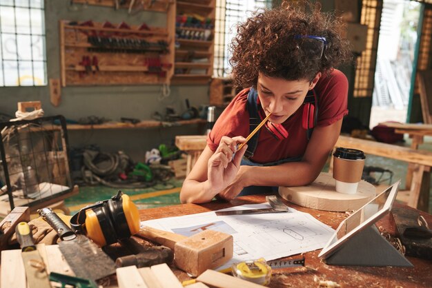 Foto jovem mulher encaracolada debruçada sobre uma mesa bagunçada e olhando pensativamente para o projeto com um novo projeto com um lápis na boca