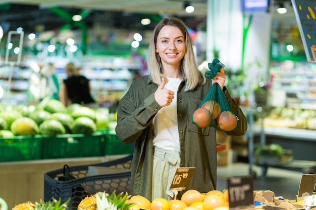 Jovem mulher escolhe e escolhe frutas ou legumes em saco ecológico Laranjas no supermercado. cliente do sexo feminino em uma mercearia perto do balcão compra e joga um pacote reutilizável no mercado