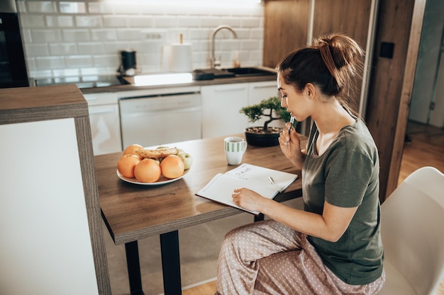 Jovem mulher escrevendo em um caderno e fazendo uma lista de tarefas enquanto saboreia o café da manhã em sua cozinha em casa.