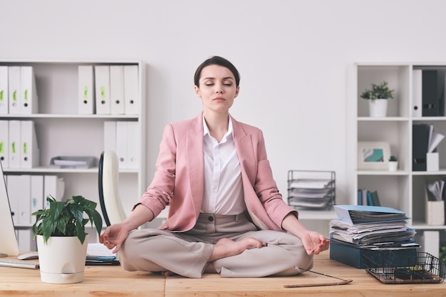 Jovem mulher espiritual com uma jaqueta rosa sentada em posição de lótus na mesa e meditando no escritório