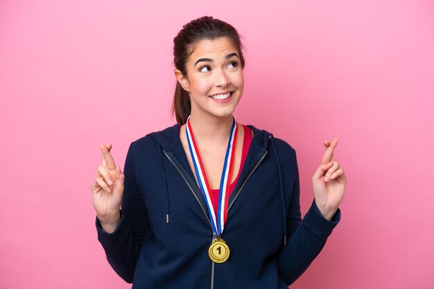 Jovem mulher esportiva brasileira com medalhas isoladas em fundo rosa com dedos cruzando