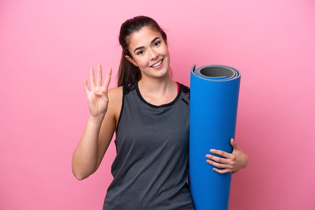 Jovem mulher esportiva brasileira indo para aulas de ioga enquanto segura um tapete isolado no fundo rosa feliz e contando quatro com os dedos