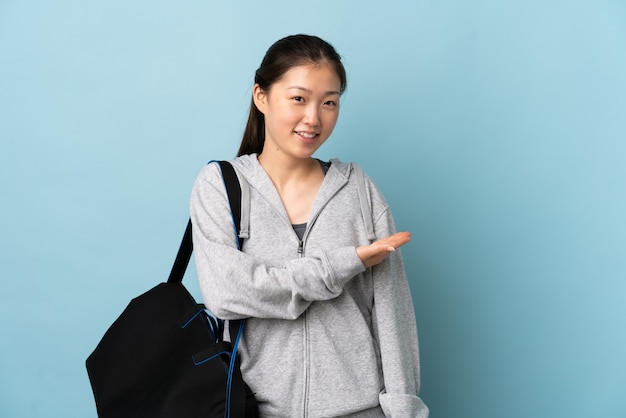 Jovem mulher esportiva chinesa com bolsa esportiva sobre uma parede azul isolada, apresentando uma ideia enquanto olha sorrindo para