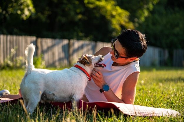 Jovem mulher esportiva, com cabelo preto curto, treinando ao ar livre em uma esteira de ioga na grama verde de um parque, atividades com o cachorro, jack russell terrier caminhada