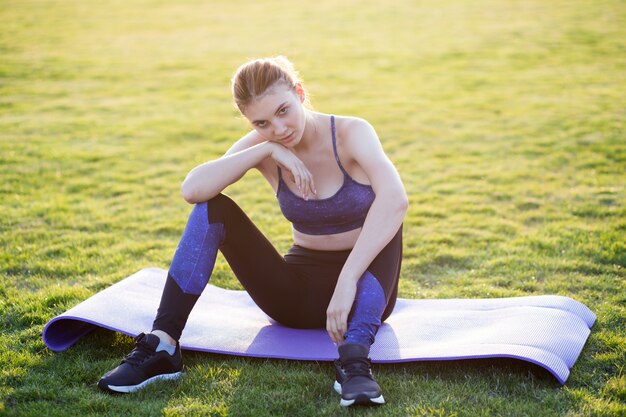 Jovem mulher esportiva em roupas esportivas, sentado no tapete de treinamento antes de fazer exercícios no campo, ao nascer do sol.