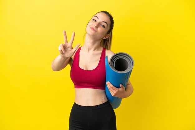 Jovem mulher esportiva indo para aulas de ioga, segurando um tapete feliz e contando três com os dedos