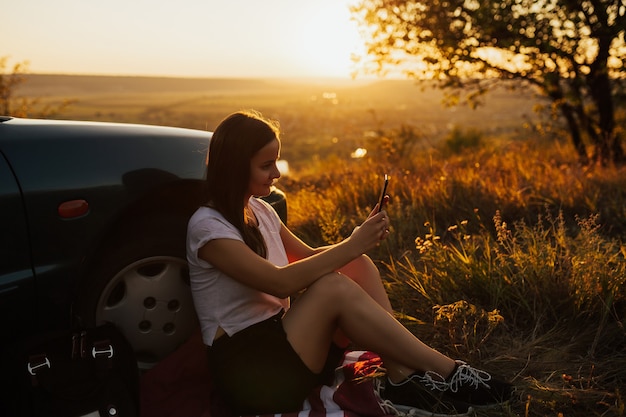 Jovem mulher está sentada perto do carro e olha para o telefone inteligente na viagem ao pôr do sol incrível.