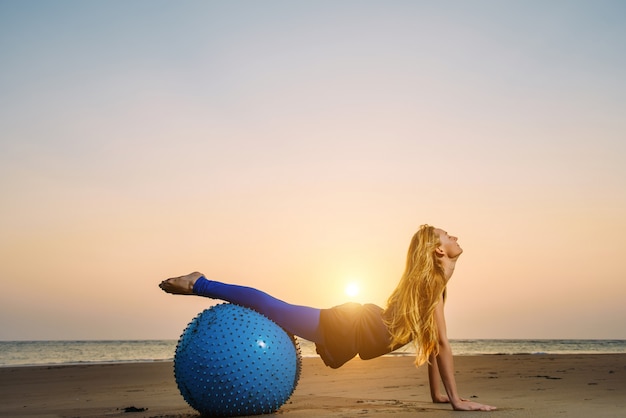 Jovem mulher, estendendo-se na bola de treinamento contra o pôr do sol sobre o mar
