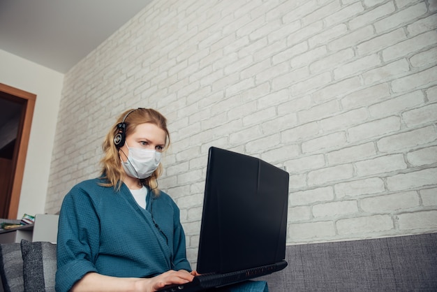 Jovem mulher européia caucasiana em máscara médica e fones de ouvido usando o laptop no sofá durante o trabalho remotamente em casa devido à pandemia de coronavírus