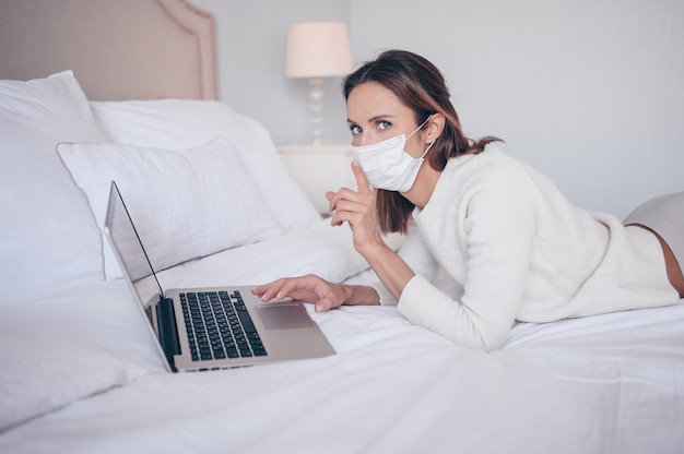 Jovem mulher européia na máscara de medicamento facial trabalhando em um laptop no quarto durante o isolamento do coronavírus