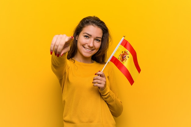 Jovem mulher europeia segurando um sorriso alegre bandeira espanhola apontando para a frente