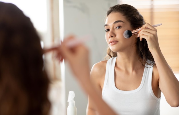 Jovem mulher fazendo maquiagem aplicando pó facial com pincel dentro de casa