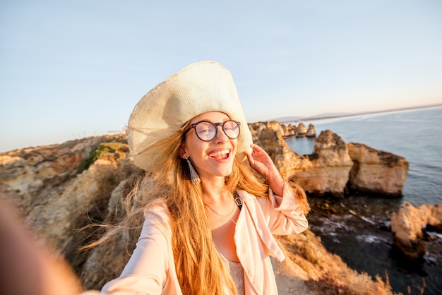 Jovem mulher fazendo uma selfie na costa rochosa durante o nascer do sol em uma viagem em Lagos, no sul de Portugal