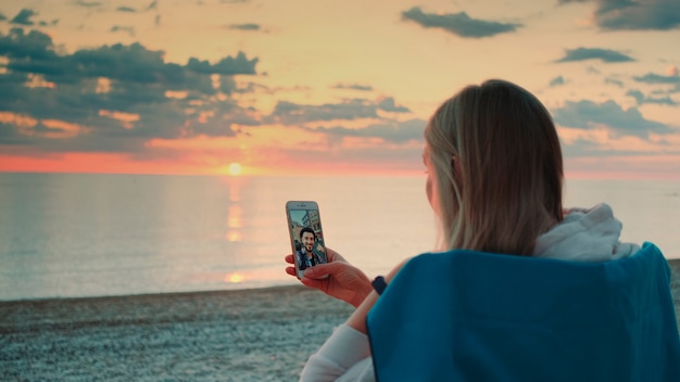 Jovem mulher fazendo videochamada para a amiga com o smartphone na praia, relaxando e falando com ...