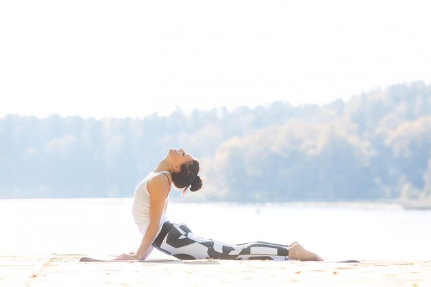 Jovem mulher fazendo yoga perto do lago ao ar livre, meditação. Esporte fitness e exercício na natureza. Pôr do sol de outono.