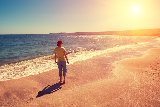 Jovem mulher feliz andando descalça na praia de volta para a câmera ao pôr do sol