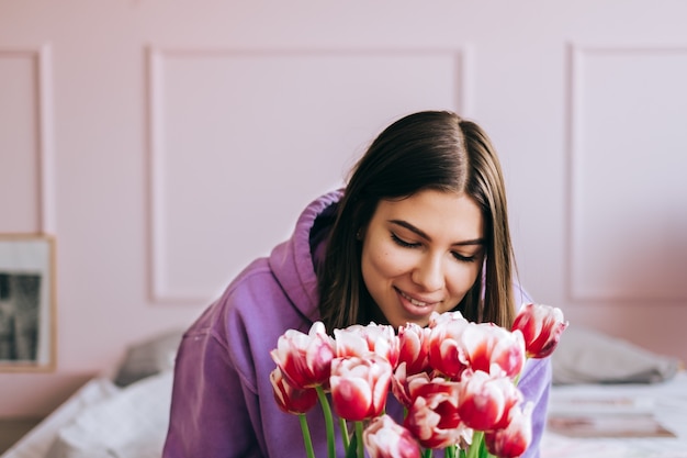 Jovem mulher feliz caucasiana cheirando o buquê de tulipas frescas em casa. sentado na cama na manhã de primavera, o conceito do dia da mulher.