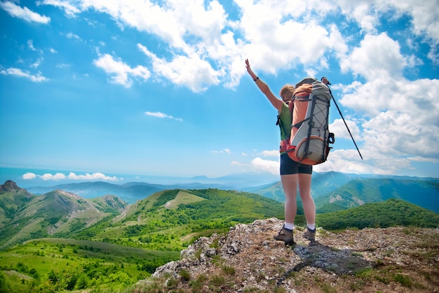 Jovem mulher feliz com um bastão de caminhada, mochila em pé na rocha com as mãos levantadas e olhando para a paisagem verde