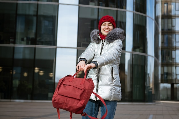 Jovem mulher feliz com um casaco quente e um chapéu vermelho, com uma mochila viaja pelas cidades