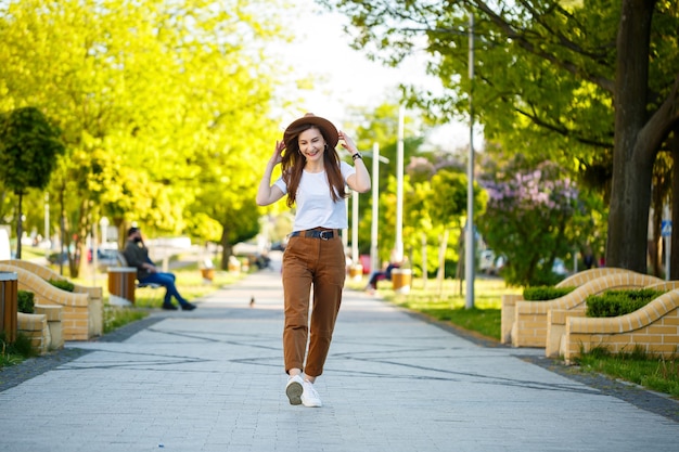 Foto jovem mulher feliz com um chapéu caminha ao longo de um beco em um parque. uma garota de aparência europeia com um sorriso no rosto em um dia ensolarado de verão