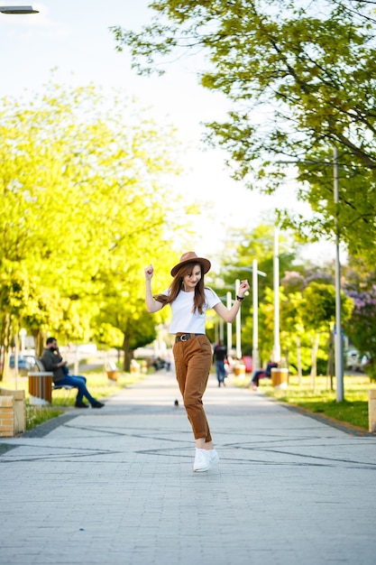 Jovem mulher feliz com um chapéu caminha ao longo de um beco em um parque. Uma garota de aparência europeia com um sorriso no rosto em um dia ensolarado de verão