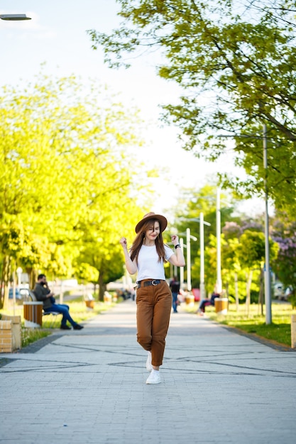 Jovem mulher feliz com um chapéu caminha ao longo de um beco em um parque. Uma garota de aparência europeia com um sorriso no rosto em um dia ensolarado de verão