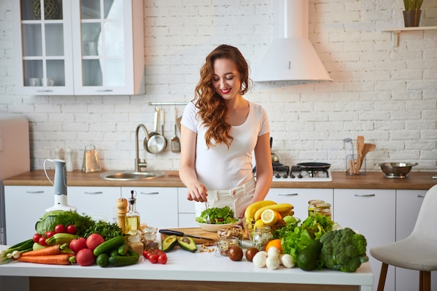 Jovem mulher feliz comendo salada na bela cozinha com ingredientes frescos verdes dentro de casa. Conceito de comida saudável