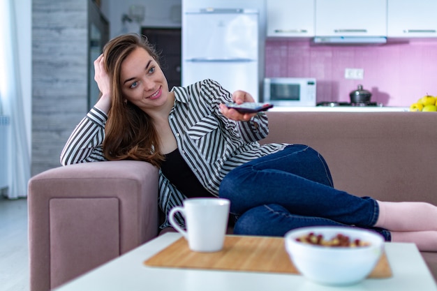 Jovem mulher feliz, descansando no sofá e assistindo tv em casa