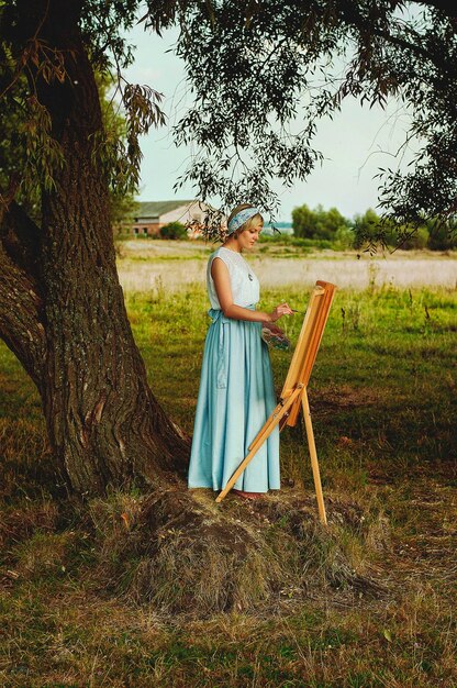 Jovem mulher feliz elegante de vestido azul pinta um quadro na natureza. Roupas vintage. Paisagem rural