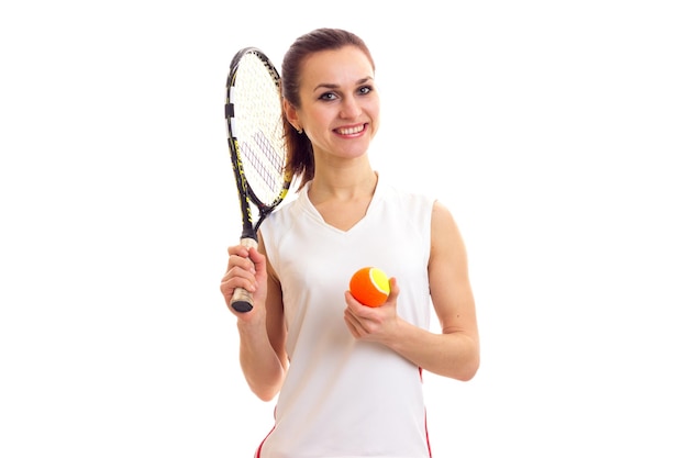 Jovem mulher feliz em camisa esportiva branca com rabo de cavalo escuro segurando raquete de tênis e bola laranja