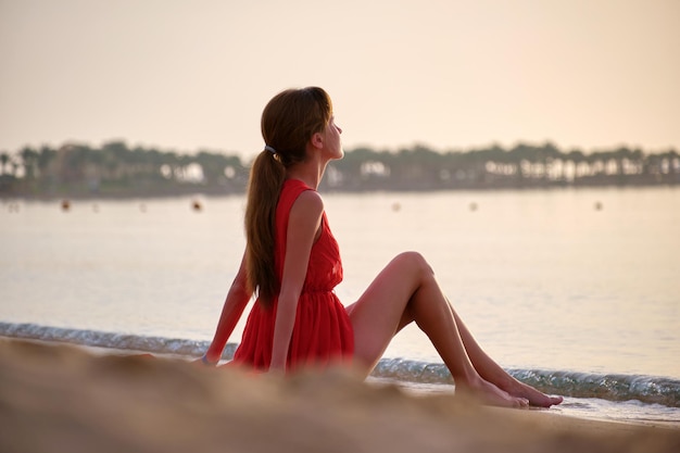 Jovem mulher feliz em um vestido vermelho relaxante na praia à beira-mar, apreciando a quente manhã tropical.