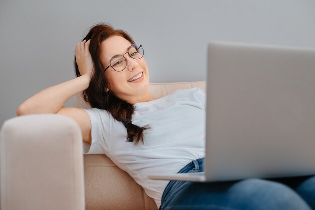 Jovem mulher feliz está descansando em casa em um sofá branco assistindo a um filme em um laptop - conceito de o ...