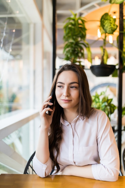 Jovem mulher feliz falando no celular com um amigo enquanto está sentada sozinha no interior de uma cafeteria moderna, sorridente garota hippie ligando para o celular enquanto relaxa após caminhar em um dia de verão