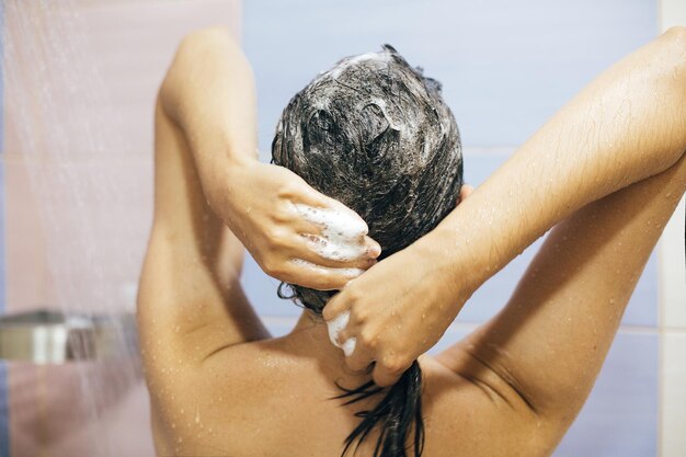 Jovem mulher feliz lavando o cabelo com xampu mãos com espuma closeup costas da linda menina morena tomando banho e aproveitando o tempo de relaxamento Conceito de estilo de vida de higiene corporal e capilar