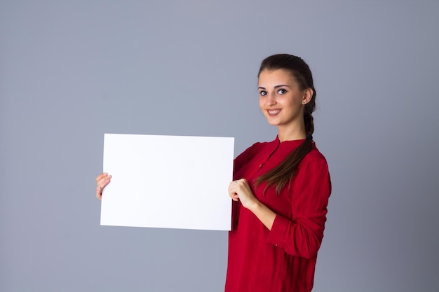Jovem mulher feliz na blusa vermelha com trança segurando a folha de papel branca sobre fundo cinza no estúdio