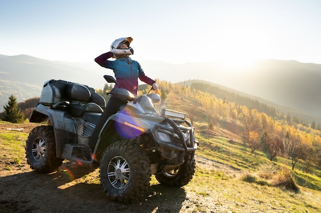 Foto jovem mulher feliz no capacete protetor, desfrutando de passeio extremo na moto atv quad nas montanhas de outono ao pôr do sol.