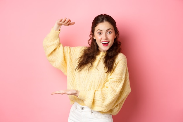 Jovem mulher feliz olhando para a câmera, mostrando um grande objeto, segurando seu logotipo ou produto com as mãos de lado no espaço da cópia, em pé contra a parede rosa.