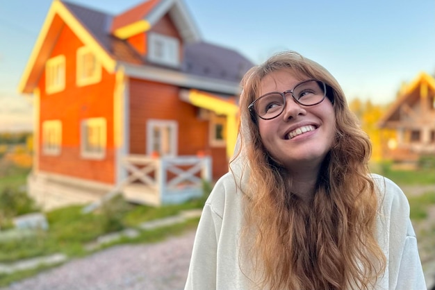 Jovem mulher feliz parada perto de sua própria casa e sorriso ao pôr do sol