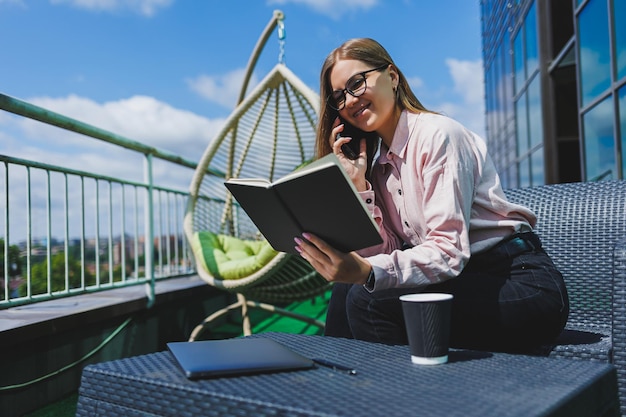 Jovem mulher feliz sorrindo e falando em uma empresária de celular fazendo anotações em um caderno enquanto relaxa em uma mesa com um netbook em um café