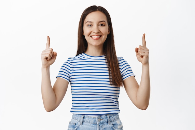 Jovem mulher feliz sorrindo, mostrando com orgulho a bandeira, apontando os dedos para cima e olhando alegre na frente, mostrar o logotipo de desconto de venda, em pé contra uma parede branca.