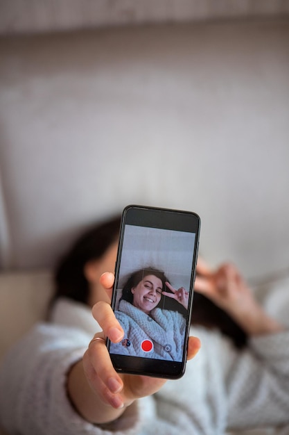 Jovem mulher feliz tomando história de selfie deitada na cama