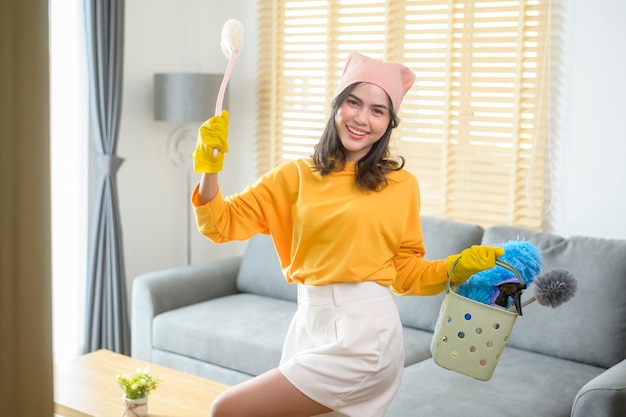 Jovem mulher feliz usando luvas amarelas e segurando uma cesta de material de limpeza na sala de estar