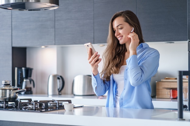 Jovem mulher feliz usando smartphone e fones de ouvido sem fio brancos para ouvir música e áudio livro na cozinha em casa. Pessoas móveis modernas