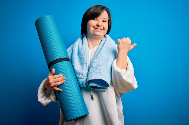 Jovem mulher fitness com síndrome de down treinando ioga e pilates segurando colchonete muito feliz apontando com a mão e o dedo para o lado