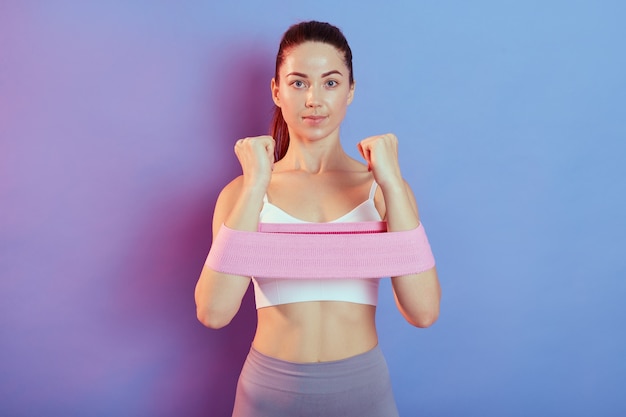 Jovem mulher forte e concentrada usando músculos de treino top branco com elástico de resistência especial, malhando, fazendo exercícios para as mãos.