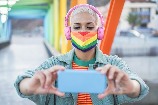 Jovem mulher gay tirando selfie ao ar livre com telefone celular - garota se divertindo com as tendências da tecnologia usando a bandeira do arco-íris - conceito lgbt