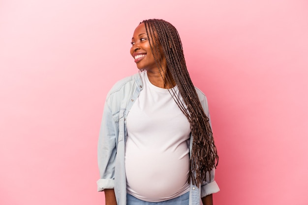 Jovem mulher grávida afro-americana isolada em um fundo rosa parece de lado sorrindo, alegre e agradável.